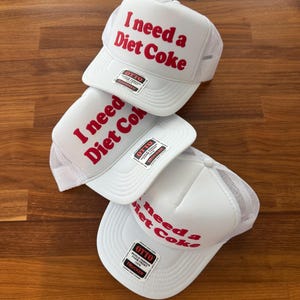May include: Three white trucker hats with mesh backs and foam fronts. Each hat features the red text "I need a Diet Coke" printed on the front panel. A black and white "OTTO" brand sticker is attached to the brim of each hat, indicating a foam front.