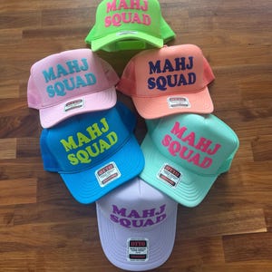 May include: A collection of six foam front trucker hats in neon green, light pink, peach, bright blue, mint green, and lavender. Each hat features the text MAHJ SQUAD in a contrasting color on the front panel and an Otto brand sticker on the brim.