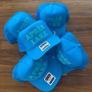 May include: A collection of blue trucker hats with the text "APR&Egrave;S LAKE" in a lighter blue font. The hats have a mesh back and a foam front. Each hat has a "OTTO" brand tag.