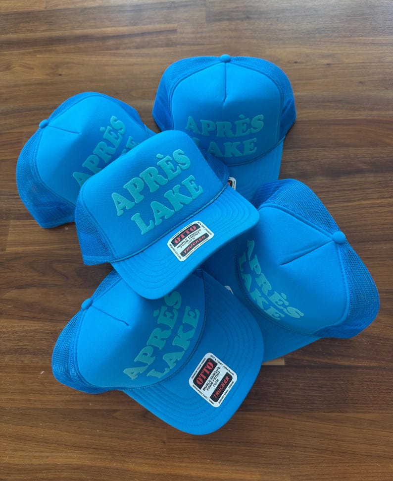 May include: A collection of blue trucker hats with the text "APR&Egrave;S LAKE" in a lighter blue font. The hats have a mesh back and a foam front. Each hat has a "OTTO" brand tag.