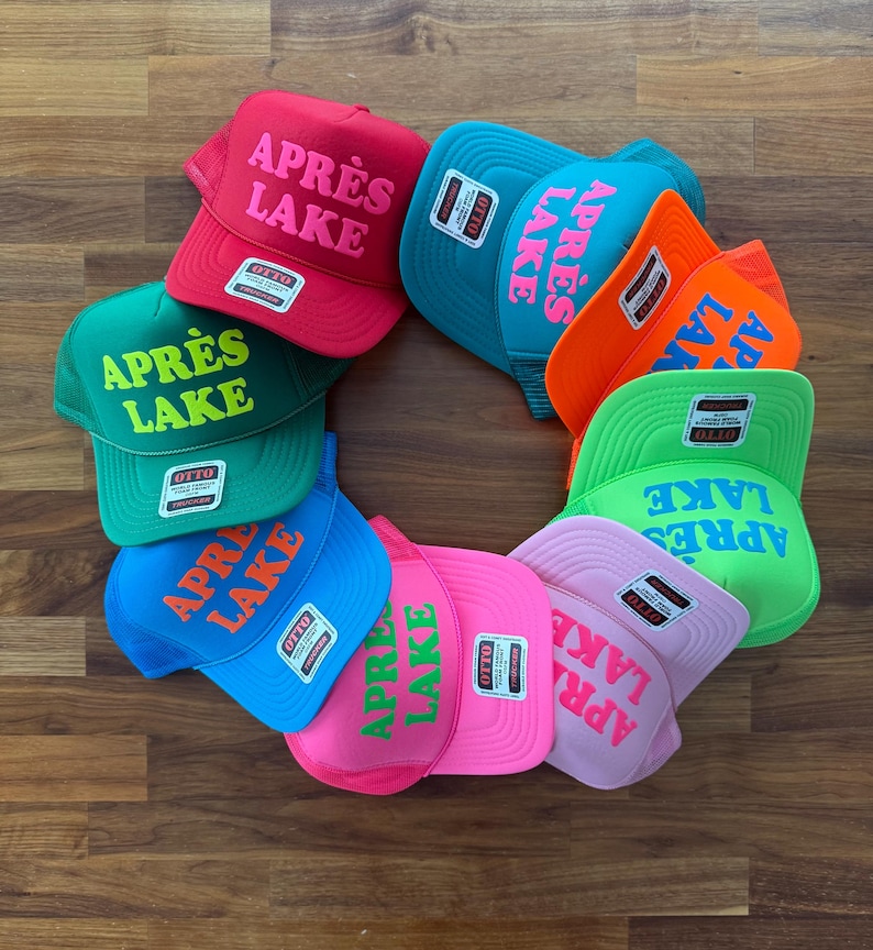 May include: A collection of ten colorful trucker hats arranged in a circle, each featuring the text APR&Egrave;S LAKE in bold lettering. The hats come in various vibrant colors including red, teal, orange, green, blue, pink, and light purple.