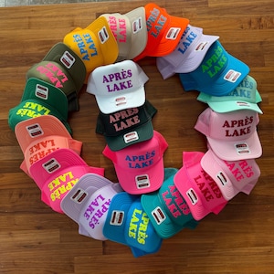 May include: A collection of colorful trucker hats arranged in a circular pattern. Each hat features the text "APR&Egrave;S LAKE" in a contrasting color. The hats come in various colors, including pink, blue, green, and orange.