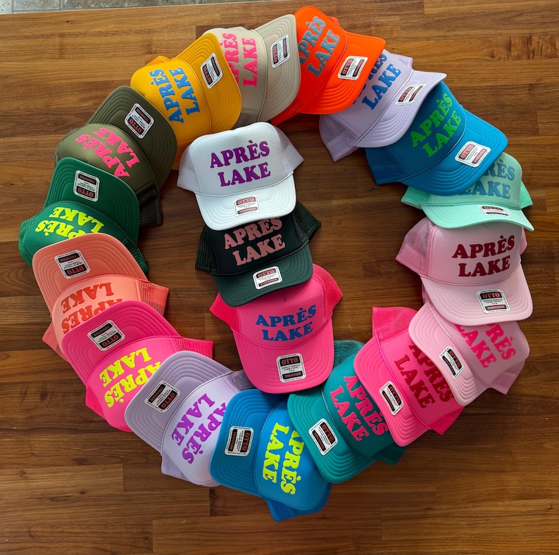May include: A collection of colorful trucker hats arranged in a circular pattern. Each hat features the text "APR&Egrave;S LAKE" in a contrasting color. The hats come in various colors, including pink, blue, green, and orange.