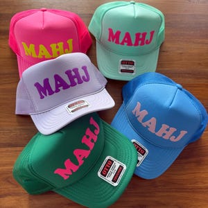 May include: Five colorful trucker hats with the word "MAHJ" in bold letters. The hats are in shades of pink, green, blue, and lavender. Each hat has a mesh back and a curved brim. The hats are displayed on a wooden surface.