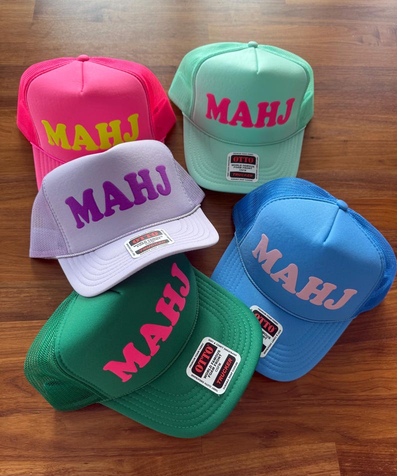 May include: Five colorful trucker hats with the word "MAHJ" in bold letters. The hats are in shades of pink, green, blue, and lavender. Each hat has a mesh back and a curved brim. The hats are displayed on a wooden surface.