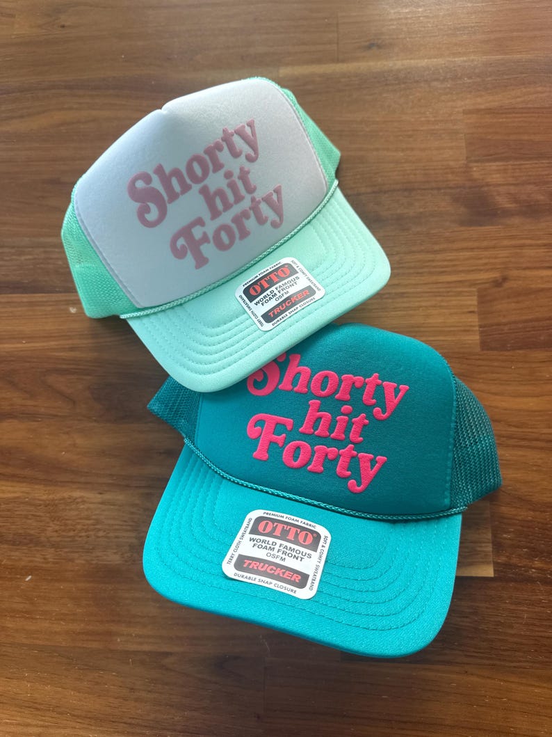 May include: Two trucker hats with the text "Shorty hit Forty" in pink script. One hat is light blue with a light green mesh, the other is teal with a dark green mesh. Both have an "OTTO" tag.
