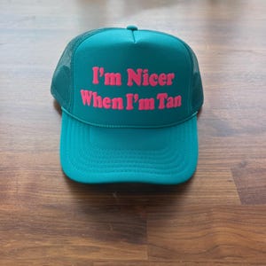 May include: A teal trucker hat featuring a mesh back and a solid front panel. The text "I'm Nicer When I'm Tan" is printed in bold, bright pink lettering across the center of the front panel. The hat has a curved brim and a matching teal button.