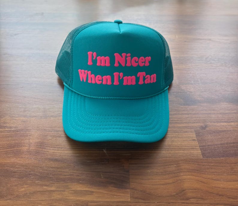 May include: A teal trucker hat featuring a mesh back and a solid front panel. The text "I'm Nicer When I'm Tan" is printed in bold, bright pink lettering across the center of the front panel. The hat has a curved brim and a matching teal button.