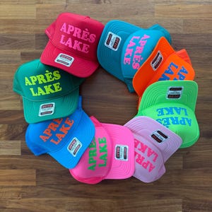 May include: A collection of colorful trucker hats arranged in a circle. Each hat features the text "APR&Egrave;S LAKE" in a contrasting color. The hats come in various colors, including red, green, blue, pink, and orange, with a mesh back.