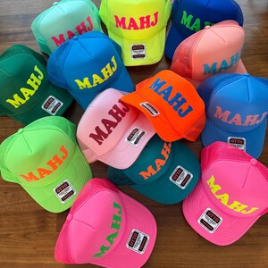 May include: A collection of colorful trucker hats with the word "MAHJ" printed across the front in various colors. The hats feature a mesh back and a curved brim. Colors include blue, pink, green, orange, and yellow.