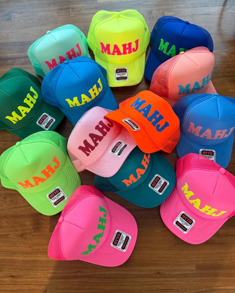 May include: A collection of colorful trucker hats with the word "MAHJ" printed across the front in various colors. The hats feature a mesh back and a curved brim. Colors include blue, pink, green, orange, and yellow.
