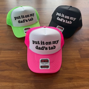May include: Three trucker hats with a white front panel and a mesh back. The front panel of each hat has black text that reads "put it on my dad's tab". The hats are in green, black, and pink.