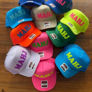 May include: A collection of trucker hats in various colors, including blue, orange, green, pink, and tan. Each hat features the word "MAHJ" in a contrasting color. The hats have a mesh back and a curved brim.