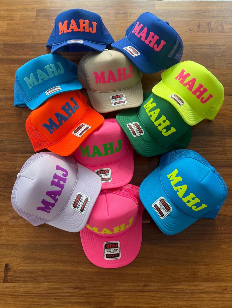 May include: A collection of trucker hats in various colors, including blue, orange, green, pink, and tan. Each hat features the word "MAHJ" in a contrasting color. The hats have a mesh back and a curved brim.