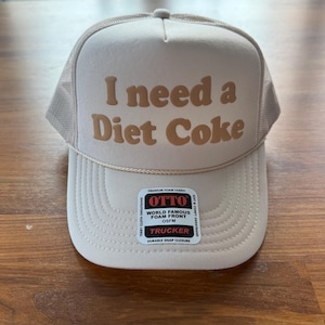 May include: A beige trucker hat featuring a mesh back and a foam front panel with the text I need a Diet Coke printed in a brown, rounded font. A decorative cord runs across the base of the crown, and a rectangular product label is attached.