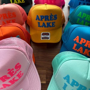 May include: A collection of trucker hats in various colors, including yellow, pink, and blue. Each hat features the text "APR&Egrave;S LAKE" in a contrasting color. The hats have a mesh back and a foam front.