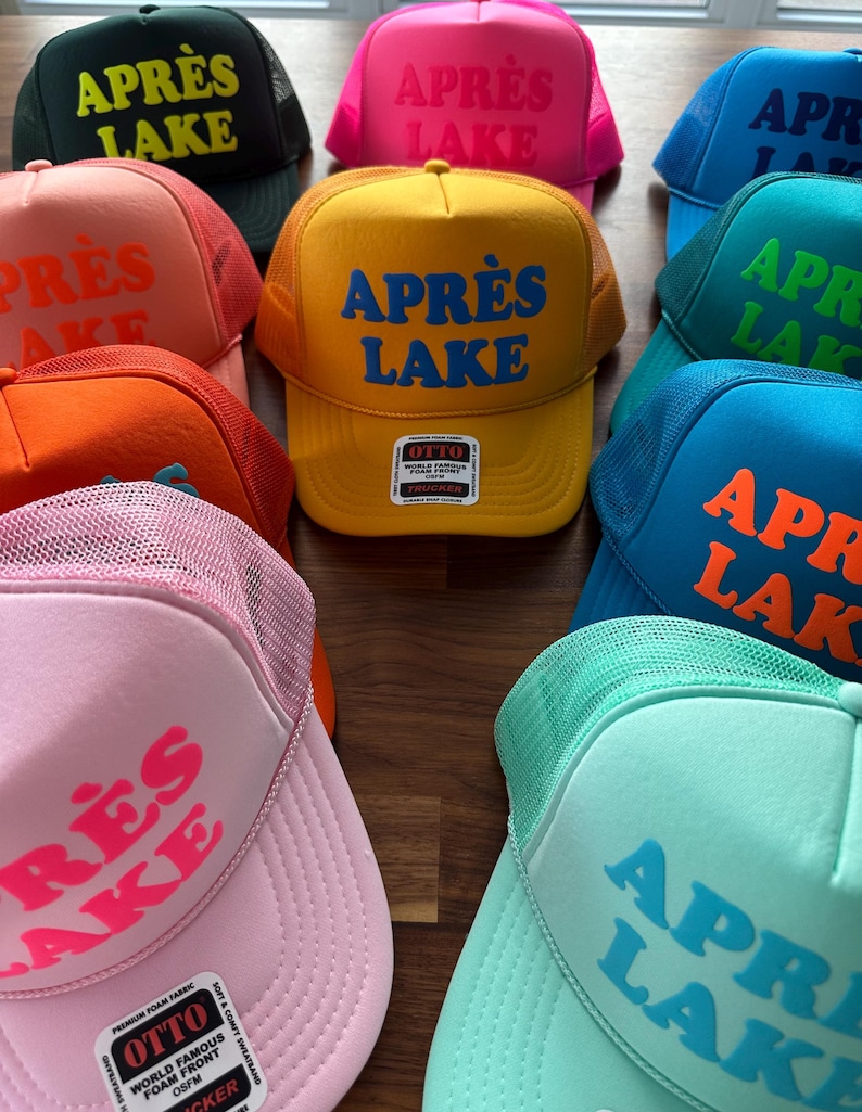 May include: A collection of trucker hats in various colors, including yellow, pink, and blue. Each hat features the text "APR&Egrave;S LAKE" in a contrasting color. The hats have a mesh back and a foam front.