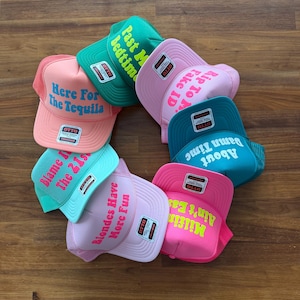 May include: A collection of seven colorful trucker hats arranged in a circle, each featuring a unique phrase in bold lettering. The hats come in shades of pink, green, and teal, with Otto brand labels on the front panels and mesh back sections.