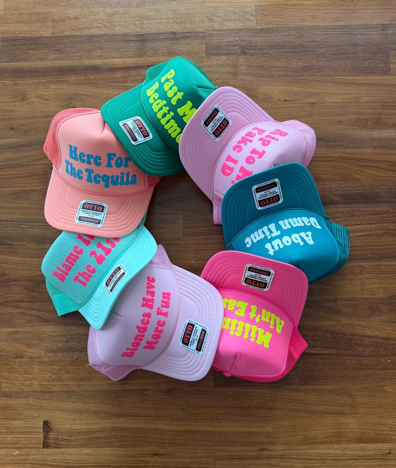 May include: A collection of seven colorful trucker hats arranged in a circle, each featuring a unique phrase in bold lettering. The hats come in shades of pink, green, and teal, with Otto brand labels on the front panels and mesh back sections.