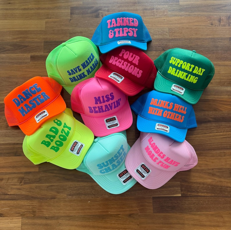May include: A collection of ten colorful trucker hats featuring various phrases including Tanned & Tipsy, Save Water Drink Margs, Pour Decisions, Support Day Drinking, Dance Master, Miss Behavin, Drinks Well With Others, Bad & Boozy, Sunset Chaser, and Blondes Have More Fun.