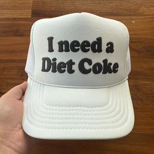 May include: A white trucker hat featuring a foam front panel with the black text I need a Diet Coke printed in a bold, rounded font. The cap includes a matching white mesh back, a structured brim with decorative stitching, and a thin white cord.