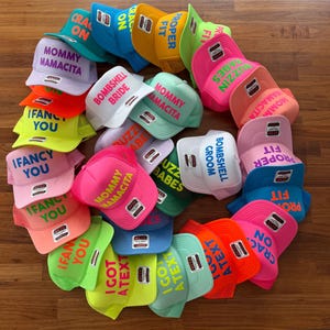 May include: A collection of colorful trucker hats with various text designs, including "Mommy Mamacita," "Bombshell Bride," and "Ifancy You." The hats are in shades of pink, orange, blue, green, and yellow, with white mesh backs.