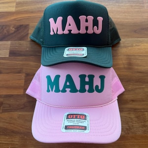 May include: Two trucker hats stacked on a wooden surface. The top hat is dark green with pink block letters spelling "MAHJ". The bottom hat is pink with green block letters spelling "MAHJ". Both hats have a mesh back and a "OTTO" label.