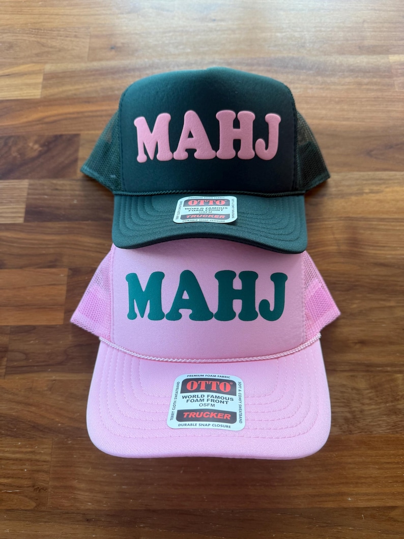May include: Two trucker hats stacked on a wooden surface. The top hat is dark green with pink block letters spelling "MAHJ". The bottom hat is pink with green block letters spelling "MAHJ". Both hats have a mesh back and a "OTTO" label.