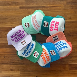 May include: A collection of six trucker hats in various colors including light purple, mint green, teal, green, blue, and peach. Each hat features a white front panel with printed text: MORE FUN THAN TWO 25 YEAR OLDS, CERTIFIED DAY DRINKER, HOT MESS, SHIT SHOW SUPERVISOR, COCKTAIL CAPTAIN, and WHISKEY MAKES ME FRISKY.