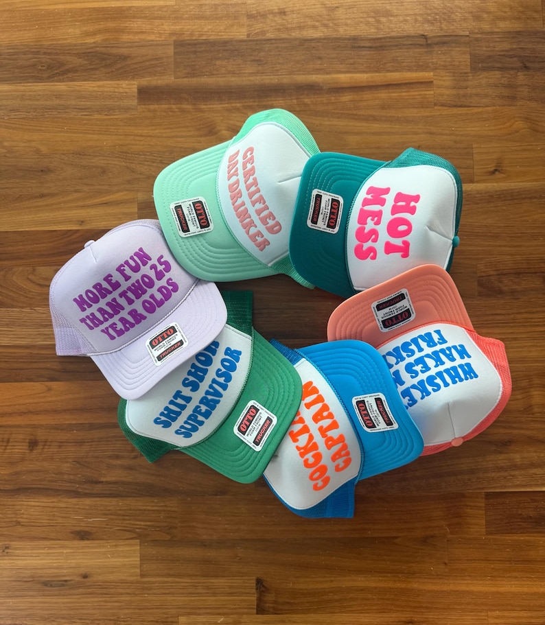 May include: A collection of six trucker hats in various colors including light purple, mint green, teal, green, blue, and peach. Each hat features a white front panel with printed text: MORE FUN THAN TWO 25 YEAR OLDS, CERTIFIED DAY DRINKER, HOT MESS, SHIT SHOW SUPERVISOR, COCKTAIL CAPTAIN, and WHISKEY MAKES ME FRISKY.