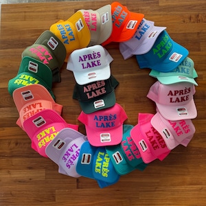 May include: A collection of colorful trucker hats arranged in a circle. Each hat features the text "APR&Egrave;S LAKE" in a contrasting color. The hats come in various colors, including pink, green, orange, and blue.
