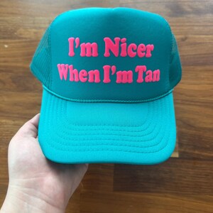May include: A teal trucker hat featuring a mesh back and a solid front panel. The text "I'm Nicer When I'm Tan" is printed in bold, bright pink lettering across the center of the front panel. The hat has a curved brim and a matching teal cord detail.