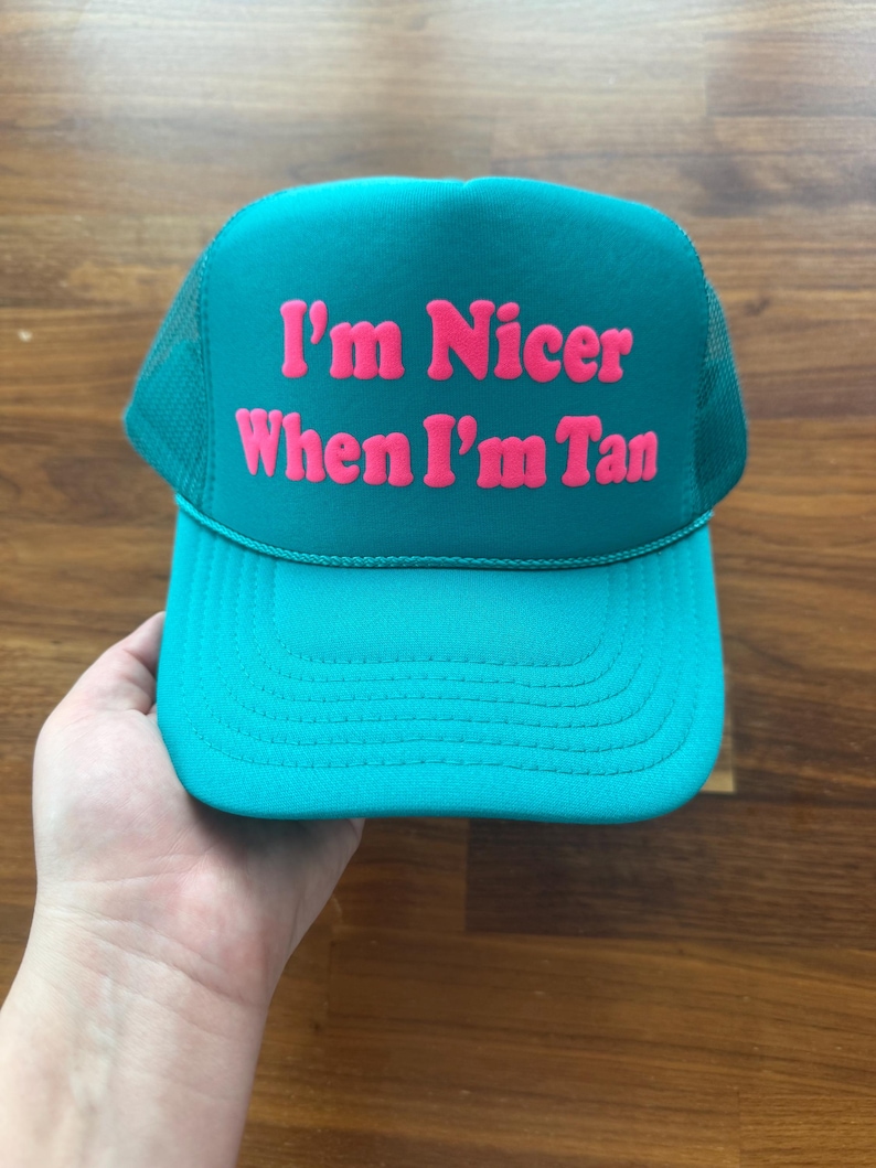 May include: A teal trucker hat featuring a mesh back and a solid front panel. The text "I'm Nicer When I'm Tan" is printed in bold, bright pink lettering across the center of the front panel. The hat has a curved brim and a matching teal cord detail.