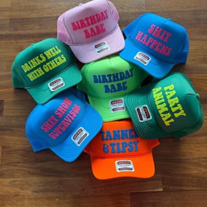May include: A collection of seven foam trucker hats in various colors including pink, blue, green, and orange. Each hat features a unique phrase printed in contrasting colors, such as Birthday Babe, Shit Happens, Drinks Well With Others, and Animal Party.