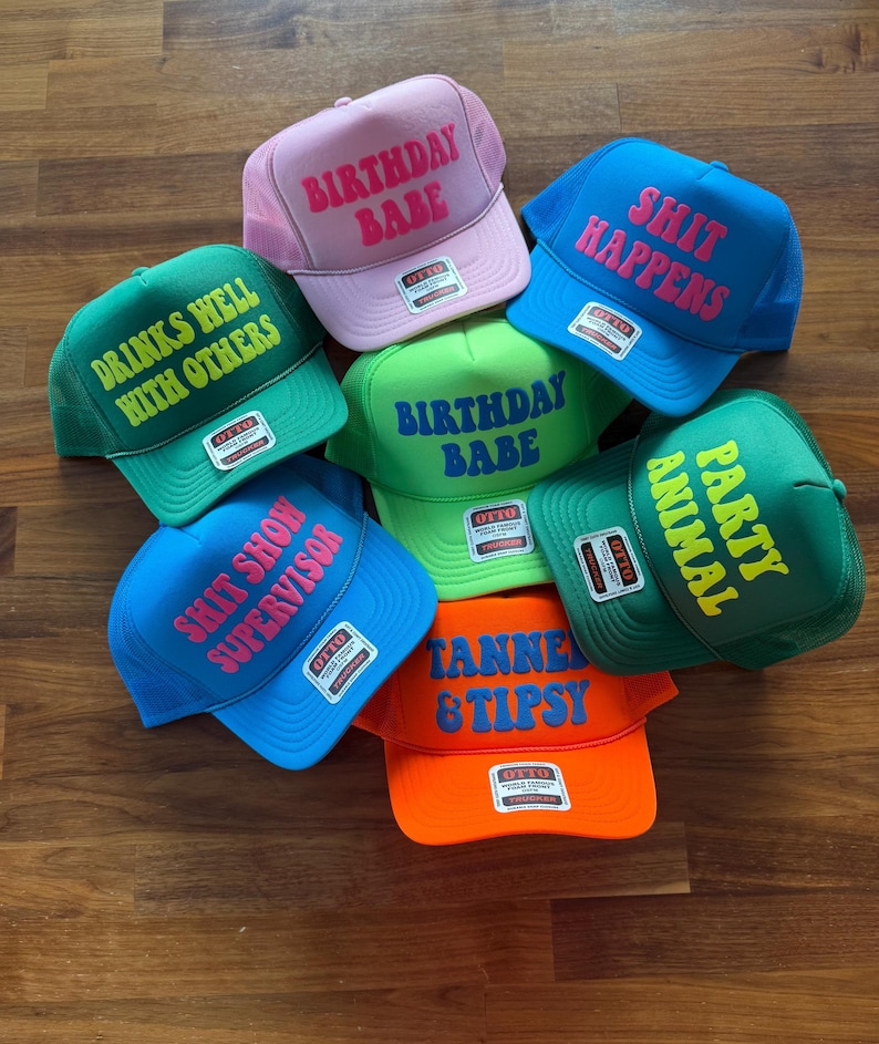 May include: A collection of seven foam trucker hats in various colors including pink, blue, green, and orange. Each hat features a unique phrase printed in contrasting colors, such as Birthday Babe, Shit Happens, Drinks Well With Others, and Animal Party.