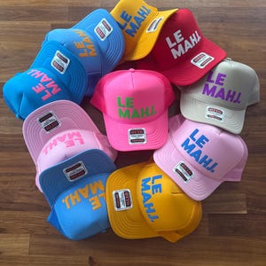 May include: A collection of ten trucker hats in various colors including yellow, red, light blue, bright blue, pink, and beige. Each hat features the text LE MAHJ. printed on the front panel in a contrasting color and includes an Otto brand label.