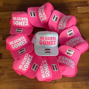 May include: A collection of trucker hats featuring a foam front panel and mesh back. Most hats are bright pink with white text reading ME GUSTA GOMEZ, while one central hat is white with bright pink text reading ME GUSTA GOMEZ. Each hat has an Otto brand label.