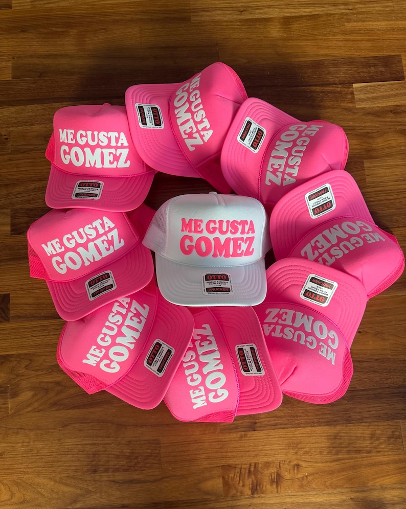 May include: A collection of trucker hats featuring a foam front panel and mesh back. Most hats are bright pink with white text reading ME GUSTA GOMEZ, while one central hat is white with bright pink text reading ME GUSTA GOMEZ. Each hat has an Otto brand label.