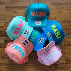 May include: A collection of six trucker hats in various colors, including teal, blue, pink, and light green. Each hat features the word "MAHJ" in a contrasting color across the front. The hats are displayed on a wooden surface.