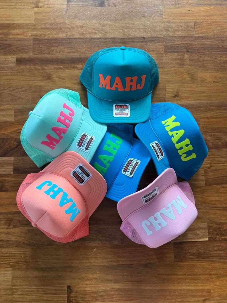 May include: A collection of six trucker hats in various colors, including teal, blue, pink, and light green. Each hat features the word "MAHJ" in a contrasting color across the front. The hats are displayed on a wooden surface.