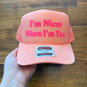 May include: A peach-colored trucker hat featuring a foam front panel with the text "I'm Nicer When I'm Tan" in bold pink lettering. The hat includes a mesh back, a matching peach brim with decorative stitching, and a small rectangular product label.