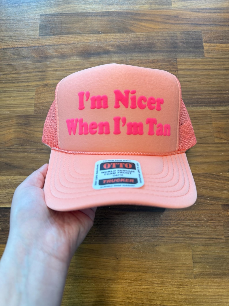 May include: A peach-colored trucker hat featuring a foam front panel with the text "I'm Nicer When I'm Tan" in bold pink lettering. The hat includes a mesh back, a matching peach brim with decorative stitching, and a small rectangular product label.