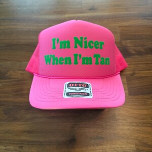 May include: A bright pink trucker hat featuring a foam front panel with the green text I'm Nicer When I'm Tan printed in a bold font. The hat includes a mesh back, a curved brim, and a white sticker on the center of the brim with OTTO branding.