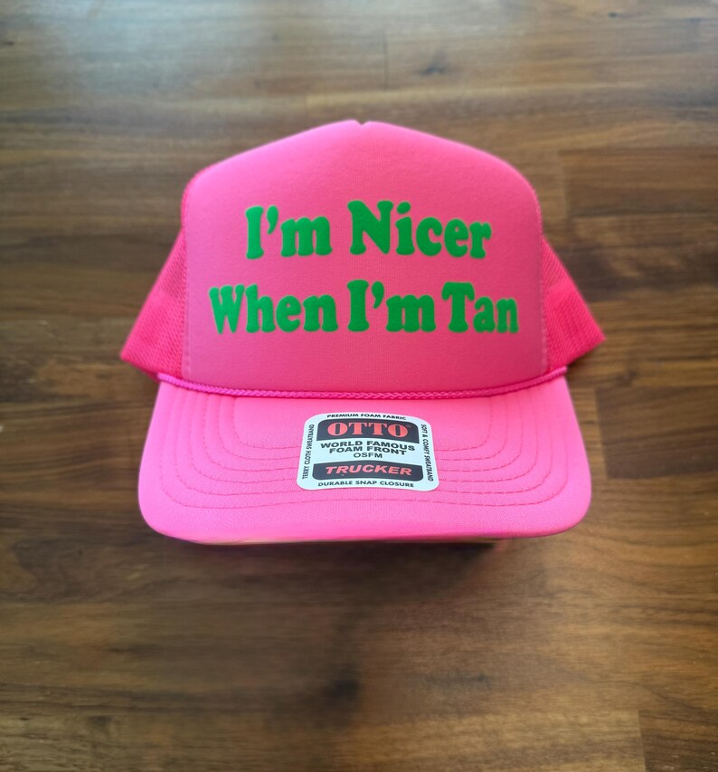 May include: A bright pink trucker hat featuring a foam front panel with the green text I'm Nicer When I'm Tan printed in a bold font. The hat includes a mesh back, a curved brim, and a white sticker on the center of the brim with OTTO branding.
