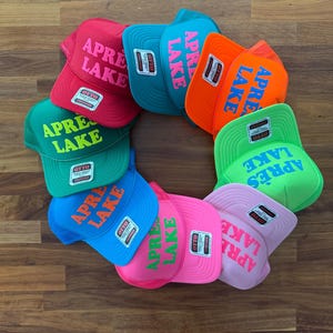 May include: A collection of colorful trucker hats arranged in a circular pattern. Each hat features the text "APR&Egrave;S LAKE" in a contrasting color. The hats come in various colors, including red, teal, orange, green, blue, pink, and light pink.