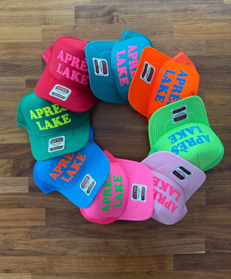 May include: A collection of colorful trucker hats arranged in a circular pattern. Each hat features the text "APR&Egrave;S LAKE" in a contrasting color. The hats come in various colors, including red, teal, orange, green, blue, pink, and light pink.