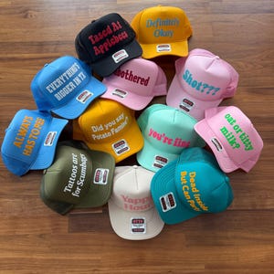 May include: A collection of eleven colorful trucker hats featuring various embroidered phrases including Tased At Applebees, Definitely Okay, EVERYTHING'S BIGGER IN TX, unbothered, Shot???, ALWAYS HAS TO PEE, Did you say Potato Famine?, you're fine, oat or titty milk?, Tattoos are Scumbags, Yappy Hour, and Dead Inside But Can Party.