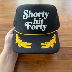 May include: Black trucker hat with the text "Shorty hit Forty" in white script. The hat features a yellow rope detail and two embroidered yellow leaf designs on the brim.