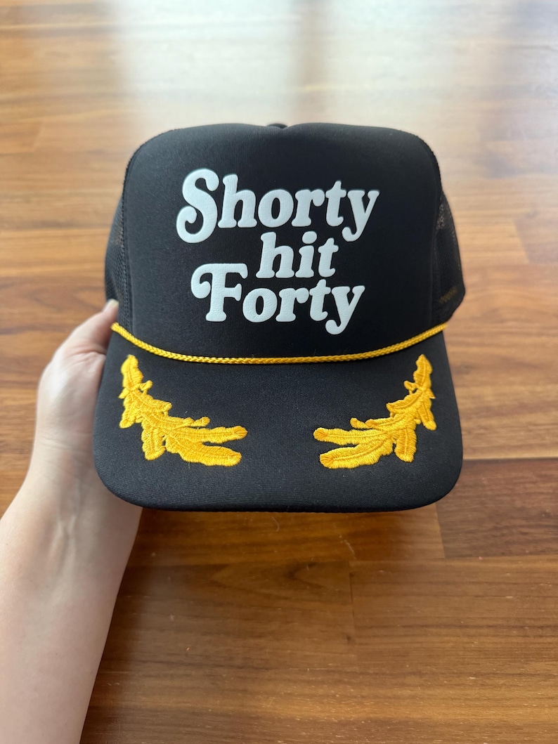 May include: Black trucker hat with the text "Shorty hit Forty" in white script. The hat features a yellow rope detail and two embroidered yellow leaf designs on the brim.