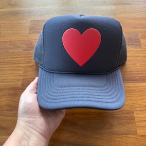 May include: A navy blue trucker hat with a large red heart in the center. The hat has a mesh back and a curved brim. The hat is a classic style, suitable for casual wear.